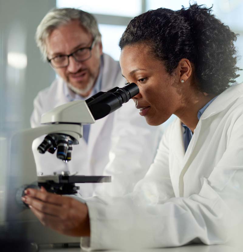 Em destaque na imagem, uma médica patologista negra, com cabelo preso em um rabo de cavalo, usando uma camisa azul com um jaleco branco por cima está olhando por um microscópio. Ao fundo, em segundo plano, um outro médico de meia idade usando óculos está observando.